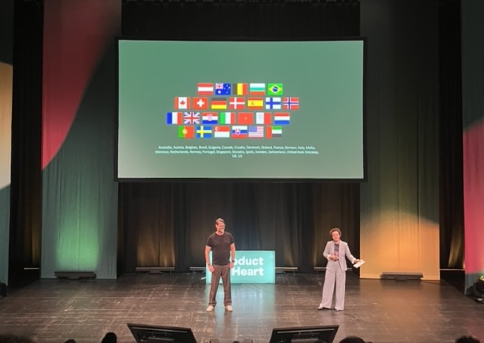 Palco do Product at Heart com um telão mostrando as bandeiras dos 27 países presentes, incluindo o Brasil